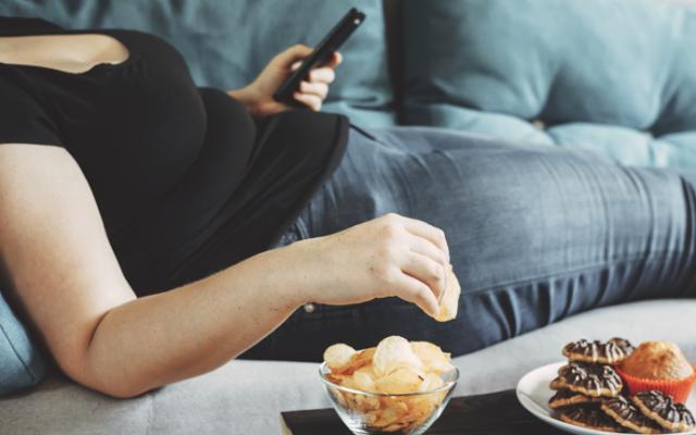 Mujer con sobrepeso mirando el móvil y comiendo comida poco sana