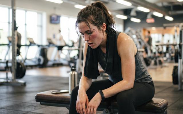 Mujer agotada sentada en el gimnasio, tras un entrenamiento intenso, evidenciando fatiga y posibles agujetas.  