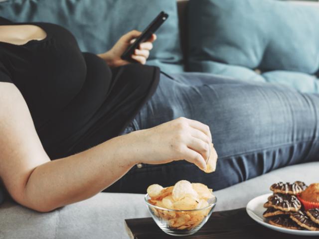 Mujer con sobrepeso mirando el móvil y comiendo comida poco sana